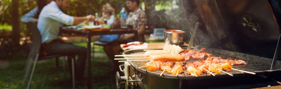 Ein voll belegter Grill vor einer Familie, die im Garten an einem Tisch isst. Auch hier gilt die Nachbarn nicht zu stören.