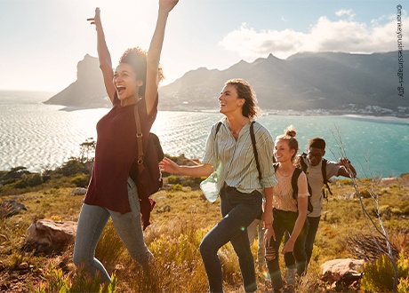 Eine Gruppe junger Menschen wandert lachend einen Hügel hinauf, mit Bergen und Meer im Hintergrund.